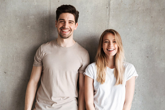 Portrait of young smiling people guy and girl 20s in basic clothing posing together at camera with happy look, isolated over concrete gray wall indoor