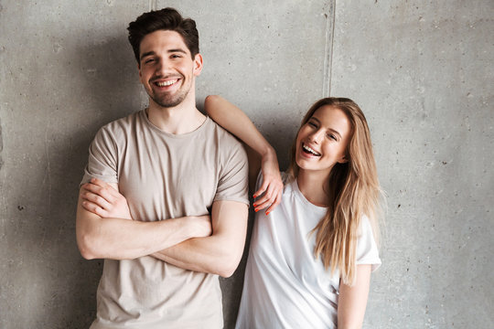 Portrait of adorable caucasian couple man and woman in basic clothing posing together at camera with happy smile, isolated over concrete gray wall indoor