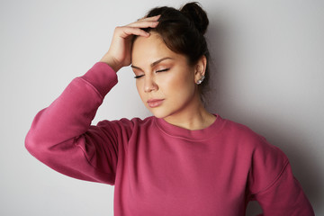 Beautiful young woman with closed eyes wearing pink hoody and holding hand on head over empty white background.Model with light nude make-up, studio background, copy paste text space.