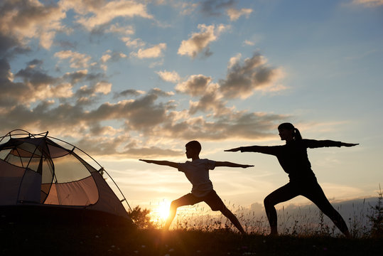Silhouette view of Yoga Warrior pose exercising by family couple in grass near tent at the daybreak on the background of morning sky with sparse clouds and bright sun. Mom grafts a son's love of sport