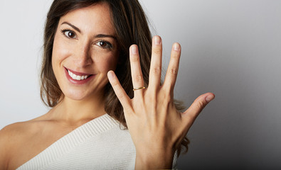 Happy european girl with long dark hair in stylish clothes smiling and holding hand on hip while showing trendy wedding ring over empty gray wall background.Copy paste text message space. Close-up