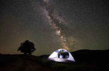 Naklejka na ściany i meble Woman traveller resting at night camping in the mountains. Rear view of girl hiker sitting inside illuminated tent, enjoying view of incredible beautiful starry sky and Milky way. Astrophotography