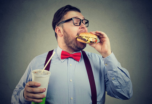 Overweight business man eating with appetite a burger holding a can of soda drink