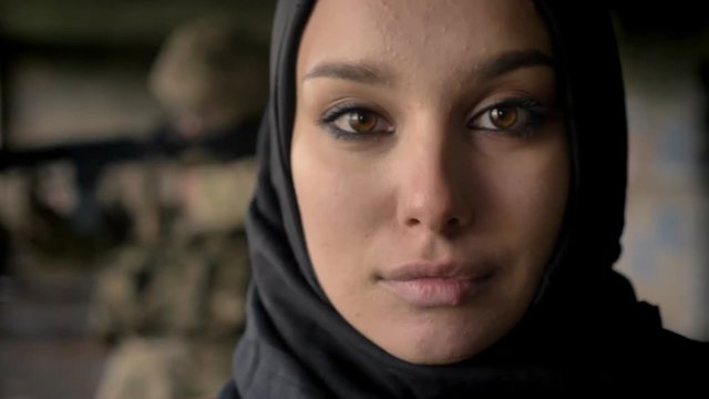 Portrait of young woman in hijab, armed soldier with weapon in background
