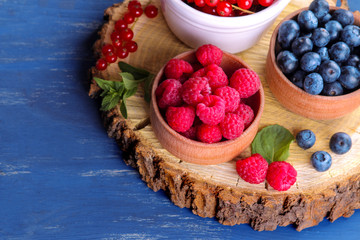 Fresh, large blueberries, currants and raspberries in bowls close-up on a wooden stand with mint stamens on a blue wooden background.
