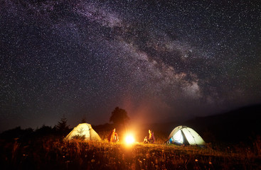 Night camping in mountains. Bright campfire burning between two backpackers, man and woman sitting...