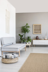 Pillow on pouf next to grey couch in scandi living room interior with plants and poster. Real photo
