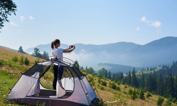 Young pretty slim girl standing on steep grassy hill at small tourist tent and taking picture of beautiful foggy mountains covered with forest under clear blue sky on bright summer morning.