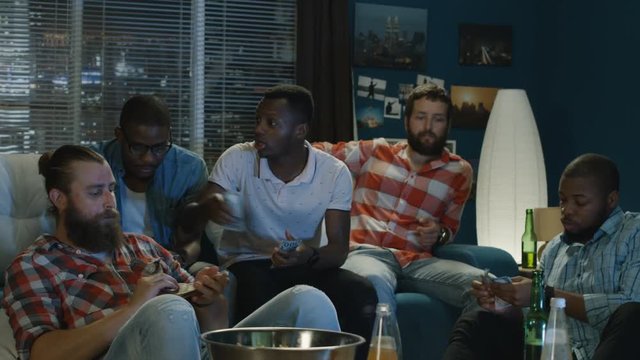 Movement shot of group of multiracial men on sofa at home making money bets for sport game while watching championship together
