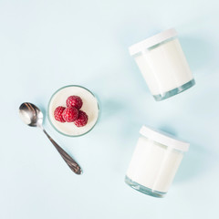 Open jar with homemade yogurt and raspberries next to closed jars on a blue background. Top view, flat lay