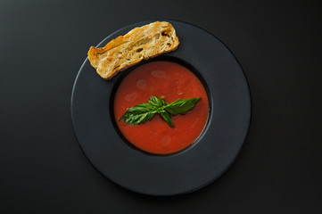 tomato soup with a toast and basil in a black plate isolated on a black background