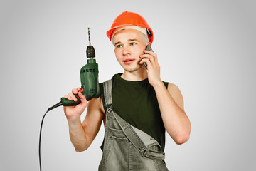 Young working guy in hard hat holds drill and calls by phone isolated on a gray background