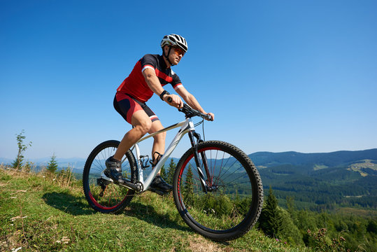 Young male sportsman cyclist in helmet, sunglasses and full equipment riding bike on grassy hill. Mountains and blue summer sky on background. Active lifestyle and extreme sport concept