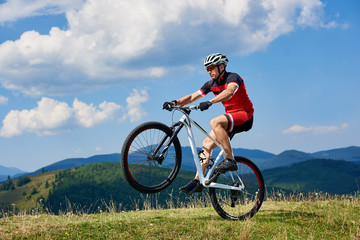Fototapeta na wymiar Athletic sportsman cyclist in professional sportswear and helmet riding cross country bike, turning on one wheel of bicycle on mountain hill. Blue summer sky on background. Extreme sport concept