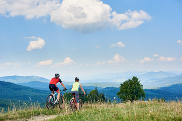 Obraz premium Rear view of active couple bikers in professional sportswear riding cross country bicycles down on the mountain road under bright blue sky with clouds on summer day in the Carpathian mountains.