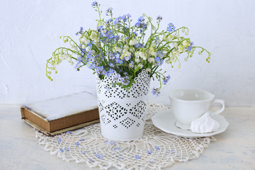 on a light table forget me nots and lilies  are placed in a vase, a book and a cup for tea and meringue
