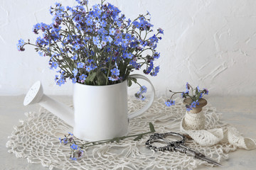 on the table of a watering can in it a bouquet from forget me nots, next there are scissors and a ribbon from a lace