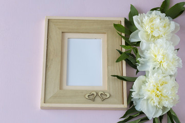 on a pink background three white peonies, an empty frame for a photo and two wooden hearts