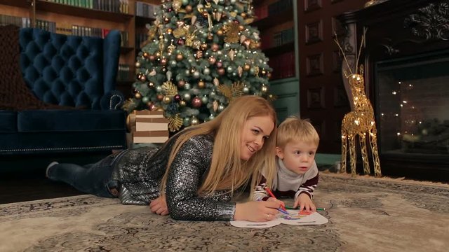 Happy mom with a little son draw lying on the floor near a decorated Christmas tree.
