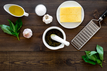 Fototapeta na wymiar Ingredients for pesto sauce. Cheese, garlic, green basil, olive oil, salt near grater and mortar on dark wooden background top view copy space
