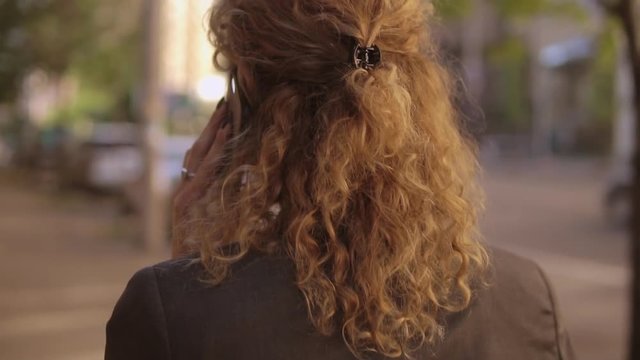 Redhaired lady speaks on celly. She walks in modern district of a big city.
