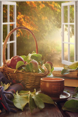 Fototapeta na wymiar Apples in a basket, a plaid book, a cup on the table near a window overlooking the autumn landscape, autumn still life, the concept of coziness in a rural house during harvesting