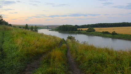 Serene landscape with dirt country road,river and field of ripe wheat.Twilight,early summer morning.