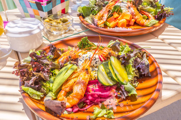 Two  mixed salads on a white wooden table with stripes caused by a sunshade in a beach bar. The healthy salad on a round plate has shrimps, avocado, cumcumber, lettuce, onion, mango and broccoli.