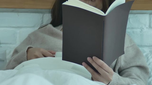 Beautiful asian woman enjoying drinking warm coffee and reading book on bed in her bedroom.Asia female wearing comfortable sweater holding a book and cup of coffee.lifestyle asia woman at home concept
