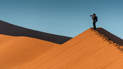 Young male traveler and photographer standing on the top of sand dune photographing sunrise or...