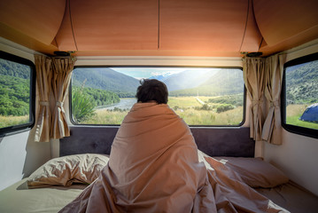 Young Asian man staying in the blanket looking at mountain scenery through the window in camper van in the morning. Road trip in summer of South Island, New Zealand. - obrazy, fototapety, plakaty