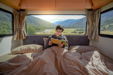 Young Asian man staying in the blanket reading magazine book in camper van with mountain scenery through the window. Road trip in summer od South Island, New Zealand.