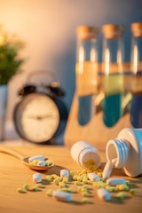 Pills spilling out of pill bottles with pharmaceutical spoon on wooden board. Table clock and test tube in the background. Prescription medicine and medical treatment concepts