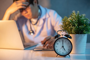 Overworked doctor working with laptop computer in hospital clinic. Male practitioner feeling stressed and tried during hard working time in medical centre. Focus on table clock.