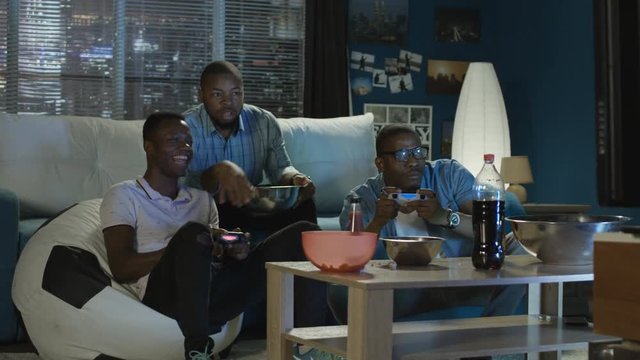 Group of young African-American men chilling on sofa with popcorn and having fun while playing videogame with gamepad and laugh together