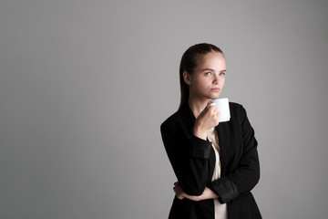 Beautiful young girl in a black jacket on a gray background holds a white cup of coffee.