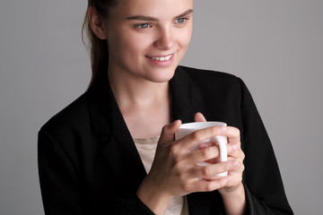 Beautiful young girl in a black jacket on a gray background holds a white cup of coffee.