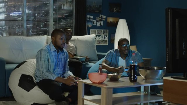 Group of young African-American men chilling on sofa with popcorn and having fun while playing videogame with gamepad one of player loses and start to laugh and tease other player his friend.