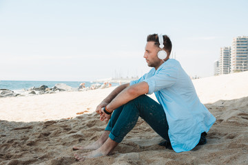 Fototapeta na wymiar Young hipster man in a blue shirt and jeans listening to music in headphones on a smartphone and is sitting on beach sand looking at sea. Digital Music lounge and relaxing concept