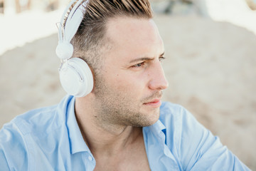 Portrait of Young hipster man in a blue shirt and jeans listening to music in headphones on a smartphone and is sitting on beach.Digital Music lounge and relaxing concept.Closeup