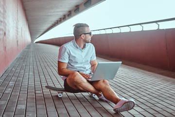 Portrait of a handsome fashionable freelancer in sunglasses dressed in a white shirt and shorts working on a laptop while sitting on a skateboard under the bridge. © Fxquadro