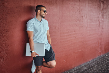 Happy handsome fashionable freelancer in sunglasses dressed in a white shirt and shorts holds a...