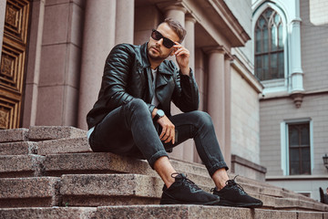 Fashionable guy dressed in a black jacket and jeans holds the smartphone sitting on steps against...