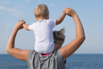 Little blonde boy sits on shoulders of his mother and watches the sea. Family vacation concept