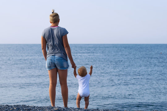 Mother And Son Together On Sea Shore. View From Behind. Mom Holds Child's Hand. The Kid Is Pointing With Another Arm At Something. Family Relationship And Learning The World Concept