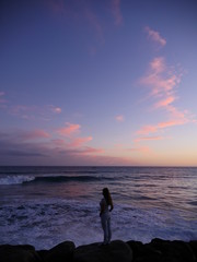 woman from behind standing on rock at the ocean shore in front a breaking wave during sunset with pink clouds