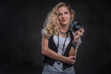 Sensual curly blonde girl photographer dressed in a white t-shirt and waistcoat wears a lot of accessories and wristwatch, posing with a camera at a studio. Isolated on a dark textured background.