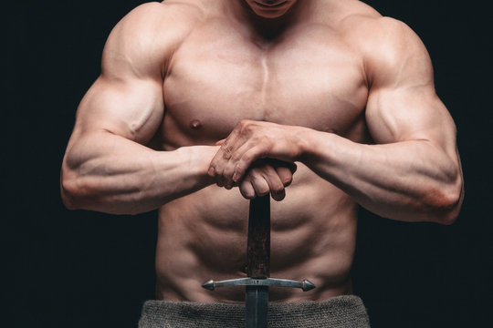 Bodybuilder man posing with a sword isolated on black background. Serious shirtless man demonstrating his mascular body. hands on a sword
