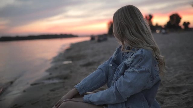 Young woman sitting on river beach at sunset looking around and dreaming