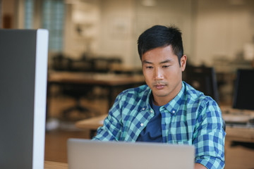 Focused young Asian designer using a laptop at his desk